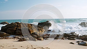 Waves crash against rocks on sandy beach. Clear blue sky, sea spray, natural coastal landscape. Untouched tropical shore