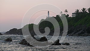 Waves crash against rocks at sunset with lighthouse on hill. Twilight ocean scene captures calming, picturesque nature