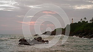 Waves crash against rocks on tropical coastline at dusk. Ocean spray, rise and fall on rough sea. Cloudy sunset sky