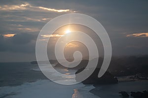 Waves crashing against rocks and sand beach at sunset with sunbeam and cloud sky