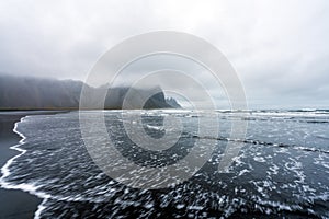 Waves are crashing ashore on Stokksnes peninsula black beach with vestrahorn mountain chain in the background.