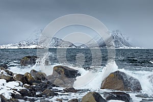 Waves crushing on the rocks on a beach from Lofoten Islands, Norway. Spectacular snowy mountains in the background. Moody winter