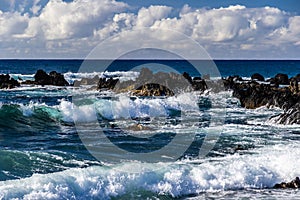 Waves on Hawiian shore; volcanic rocks in surf. Pacific in distance, with blue sky and clouds.