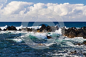 Waves on Hawiian shore; volcanic rocks in surf. Pacific in distance, with blue sky and clouds.