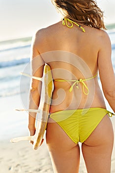 The waves look great. A slim young blonde woman standing on the beach with her surfboard on a sunny day.