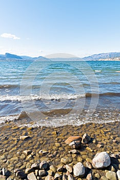 Waves on rocky shoreline of Okanagan Lake with view of blue sky and distant mountains