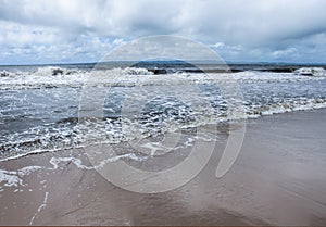 Waves rolling onto the beach with mountains on the horizon across the water and a stormy sky