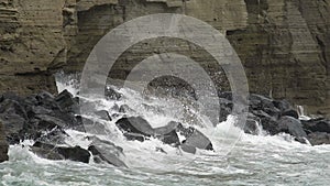 Waves of stormy sea breaking into splashes against big rock by cliff, gloomy day