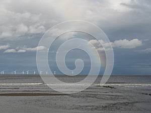 Waves washing on the shore with a beautiful rainbow above and wind turbines on the horizon
