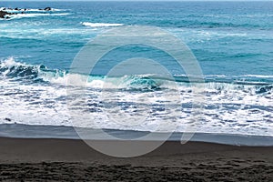 Waves with white foam on the crests of the sea with blue water and black sand
