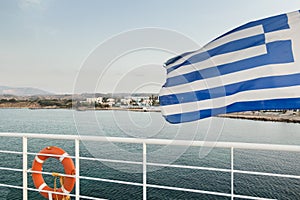 Waving Greek flag and orange lifebuoy hanging on white metal safety rail at Kos island view background
