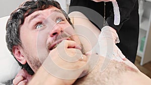 Waxing procedure - bearded adult man biting his hand while depilating hairs from his armpits in the salon