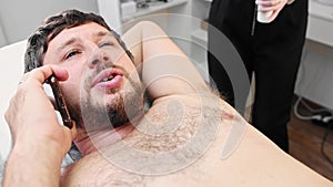 Waxing procedure - bearded man talking on his phone while depilating hairs from his armpits in the salon