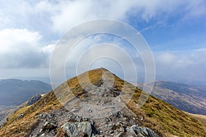 On the way to the summit trig point of a Scottish mountain Ben Vorlich with rocky path and dry grass under a majestic blue sky a
