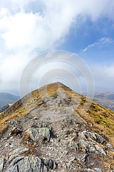 On the way to the summit trig point of a Scottish mountain Ben Vorlich with rocky path and dry grass under a majestic blue sky a