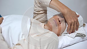 Weak elderly woman with compress on forehead lying in hospital bed, illness