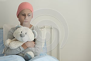 Weak girl with cancer wearing pink headscarf and hugging teddy bear next to copy space