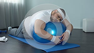 Weak-willed fat man relaxing on fitness ball, home workout break, laziness