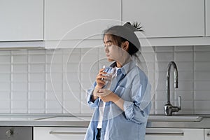 Weakened lonely Asian woman standing in kitchen of own home with glass in hands with eyes closed