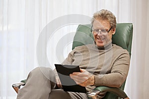 Wealthy mature man reading e-book while sitting in a chair in a bright living room