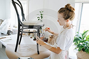 She is wearing an apron repairing an antique chair in a home white studio