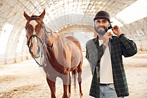 Wearing protective hat and smiling. Young man with a horse is in the hangar