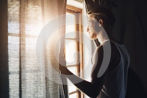 The weather looks perfect today. a handsome young man looking out of a window in his home.