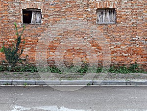 Weathered brick wall with worn windows, sidewalk with weeds and an asphalt road.