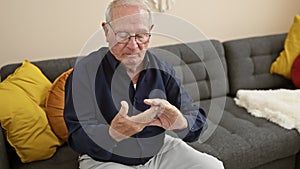 Weathered man with white hair stretching achy hand, looking serious while relaxing on room\'s sofa, suffering from arthritis at