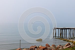 A weathered pier juts into the placid waters of Lake Garda, while a solitary boat on the horizon drifts through the fog on a quiet