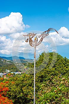 Weathervane forged cockerel on a pole against the backdrop of a tropical vertical landscape
