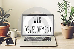 web development text on laptop screen on wooden desktop with phone, notebook, coffee and plant. web design online technology.