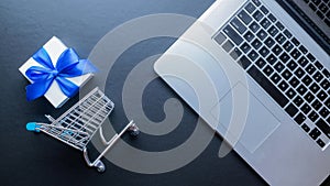 Web shopping. Laptop computer, shopping trolley and white gift with blue ribbon on dark background. Ecommerce and