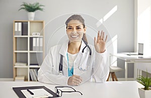 Webcam portrait of happy female online doctor sitting at desk, smiling and waving hello