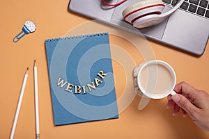 Webinar text from wooden letters on notepad on desktop with laptop and coffee cup in female hand on colored background