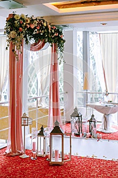 Wedding arch draped with white and pink fabric, white and red flowers on the top, decorative lamps with candles on the sides