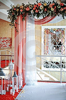 Wedding arch draped with white and pink fabric, white and red flowers on the top, decorative lamps with candles on the sides