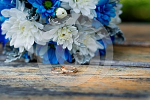 A wedding bouquet of blue and white flowers and two golden rings lie on a wooden surface. Celebration of love.