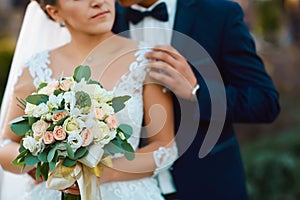 Wedding bouquet with delicate pink, beige, white flowers with green leaves in the hands of the brides in dress and costume, couple