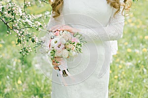 Wedding bouquet of different flowers in the hands of the bride on a green background. Bride`s hands and dress