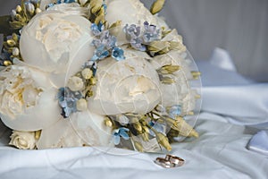 Wedding bouquet of peonies and blue flowers with 2 gold wedding rings on a white