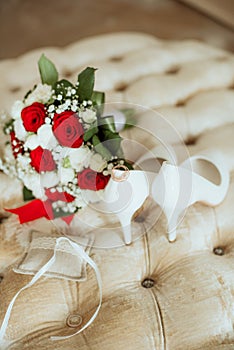 Wedding bouquet of white and red roses, white high heels and gold wedding rings on a boudoir ottoman.