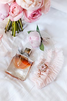 Wedding bride boutonniere, bouquet, perfume and garter laying on white bed linen. Pink bridal flowers and accessories