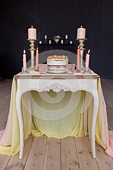 Wedding cake, candles in candlesticks and two glasses stand on a white vintage table decorated with a cloth