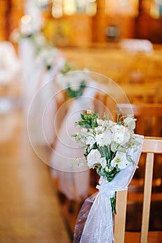 Wedding ceremony in church. Wooden chairs decorated with white tender flowers and fabric.