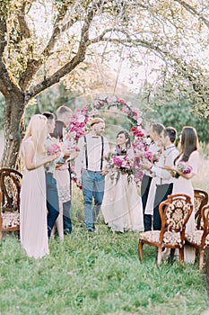 The wedding ceremony in the sunny park. The newlyweds are walking while their guests are applauding.