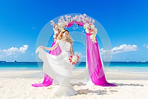 Wedding ceremony on a tropical beach in purple. Happy blond bride with wedding bouquet under the arch decorated with flowers on t