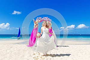 Wedding ceremony on a tropical beach in purple. Happy blond bride with wedding bouquet under the arch decorated with flowers on t