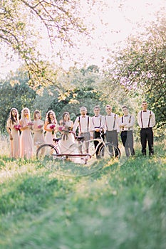 The wedding composition if the newlyweds, bridesmaids and best men behind the white bicycle in the middle of the sunny
