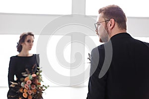 Wedding couple groom and bride posing in a white Studio. Black suit and dress. Man standing back to camera